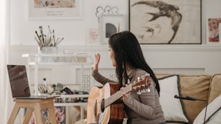 A musician playing a guitar in a cozy room with art supplies and a laptop in the background